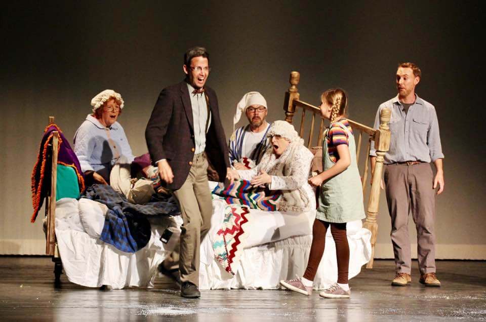An ensemble scene played around a bed on stage.